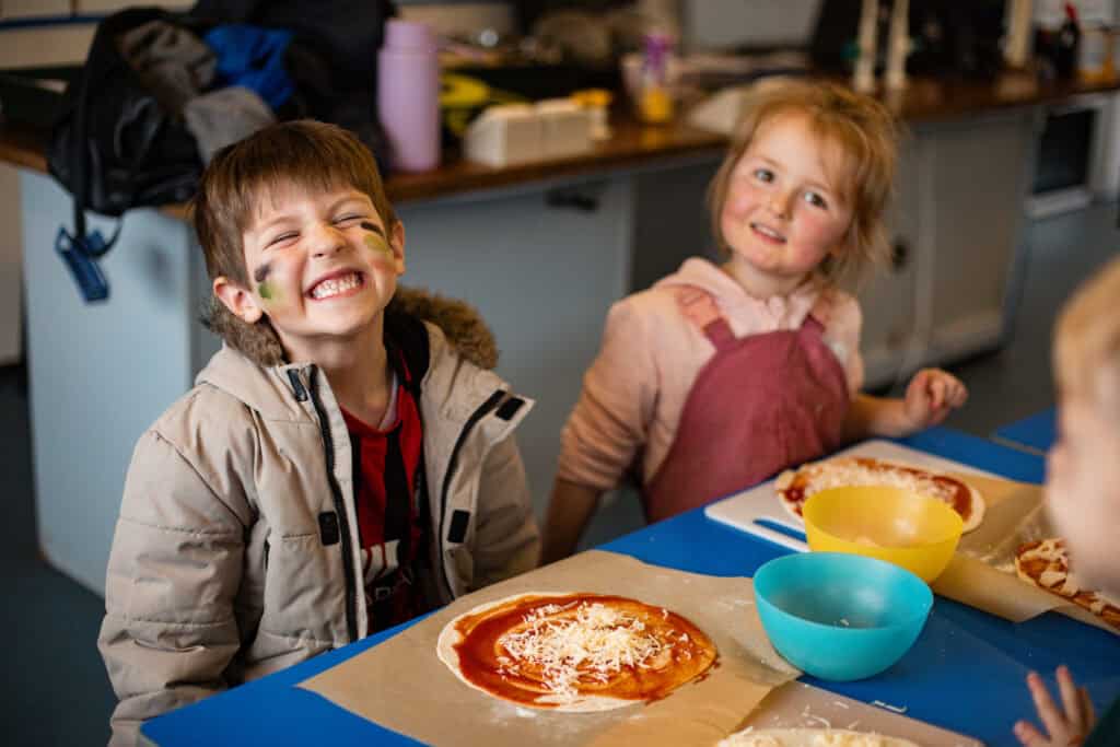 A girl and a boy smiling at the camera after adding the cheese topping to their homemade pizzas. A tasty activity.